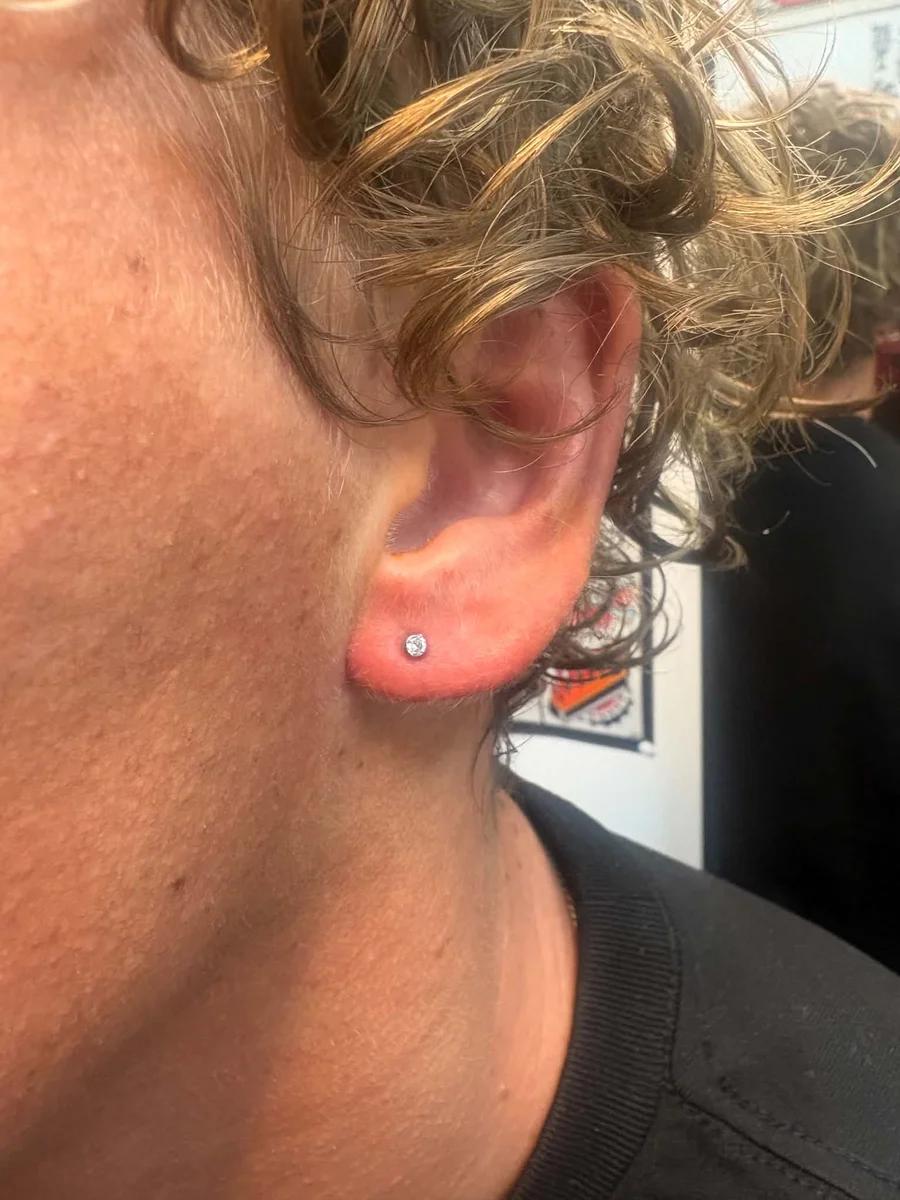 Fresh ear lobe piercing with a small crystal stud, curly hair tucked behind the ear