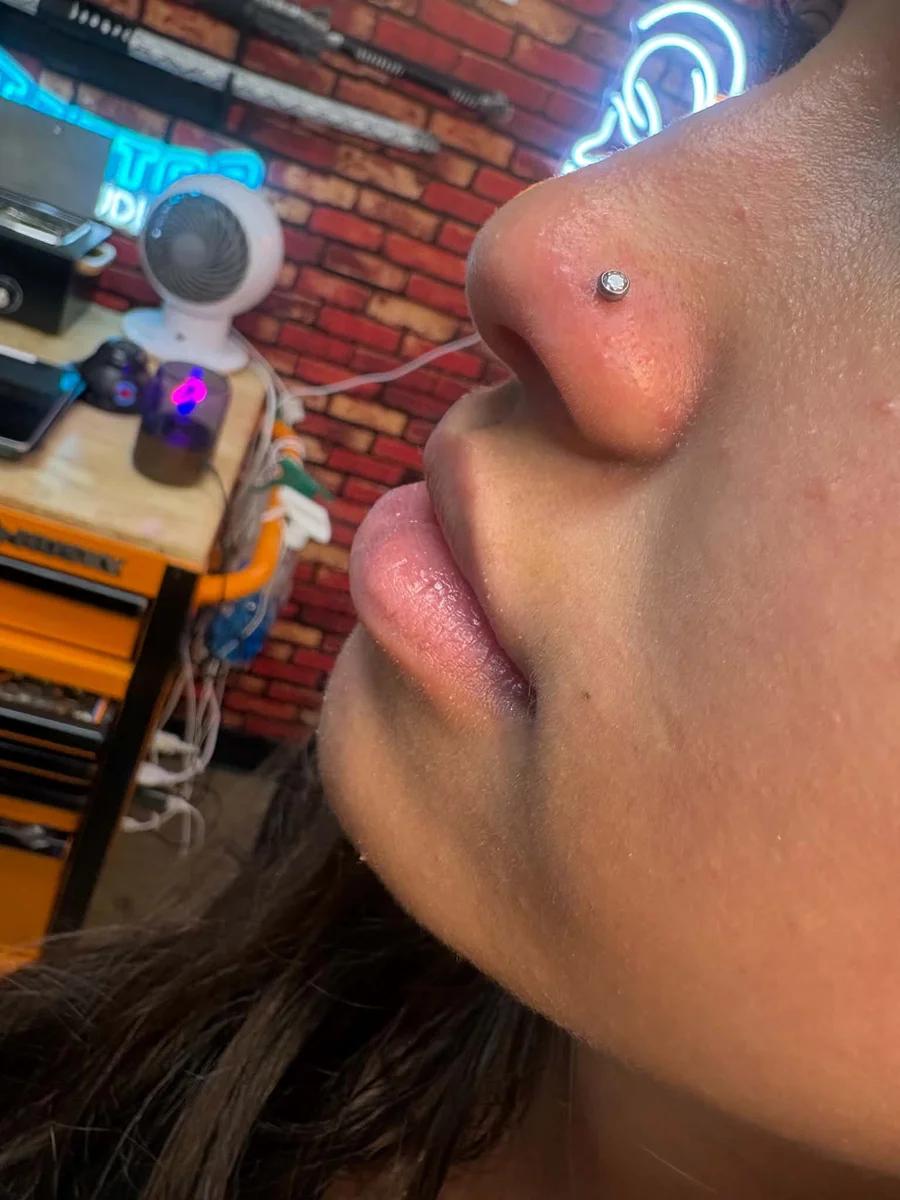 Nostril piercing with a bezel-set crystal stud, shot from below in the glow of the shop neon